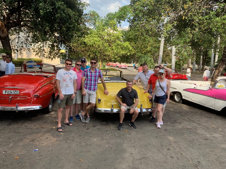 Eight men and women pose with a 1950s-era Chevrolet on the streets of Cuba.