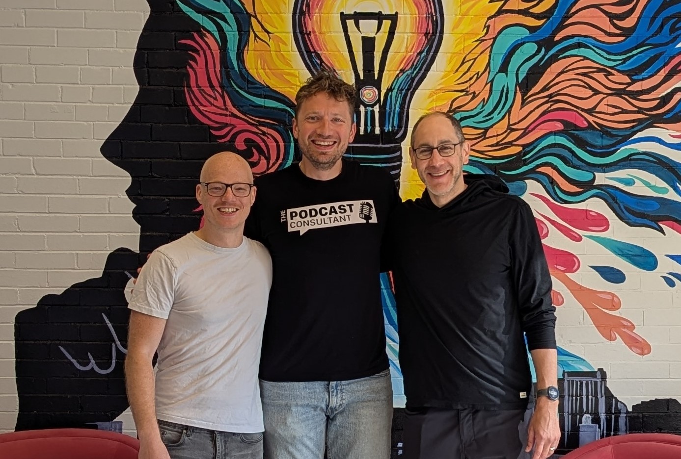 Three men in casual clothes pose in front of a colorful mural.