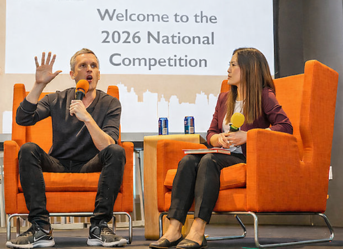 A man and woman sit in orange chairs on a stage welcoming competitors to a national competition.
