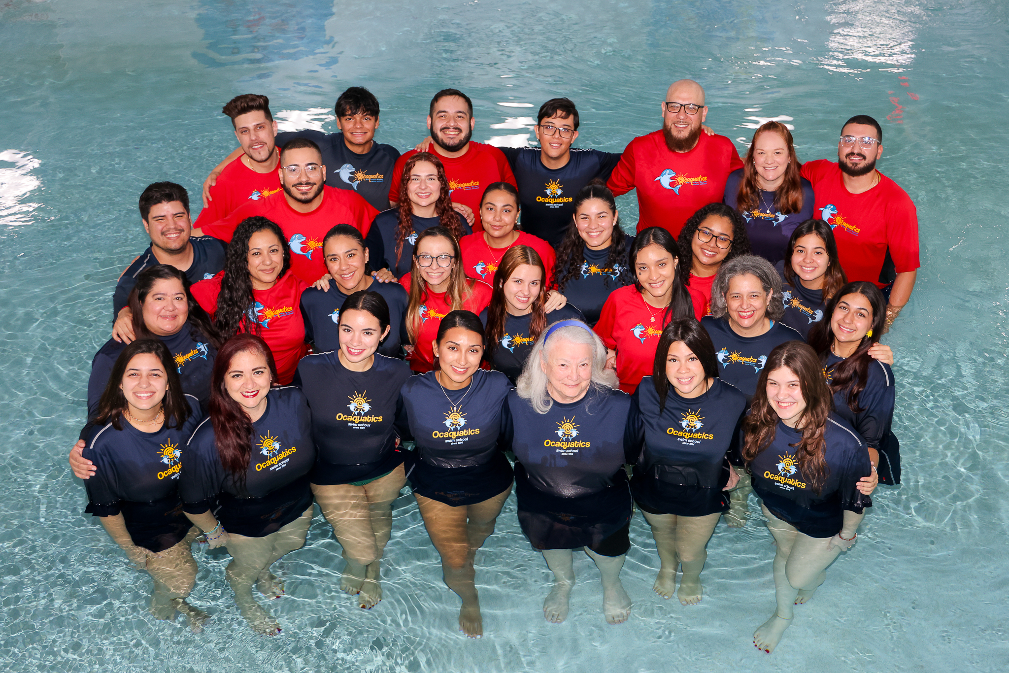 A team of about 30 swim instructors pose wearing Ocaquatics swim shirts in a pool.
