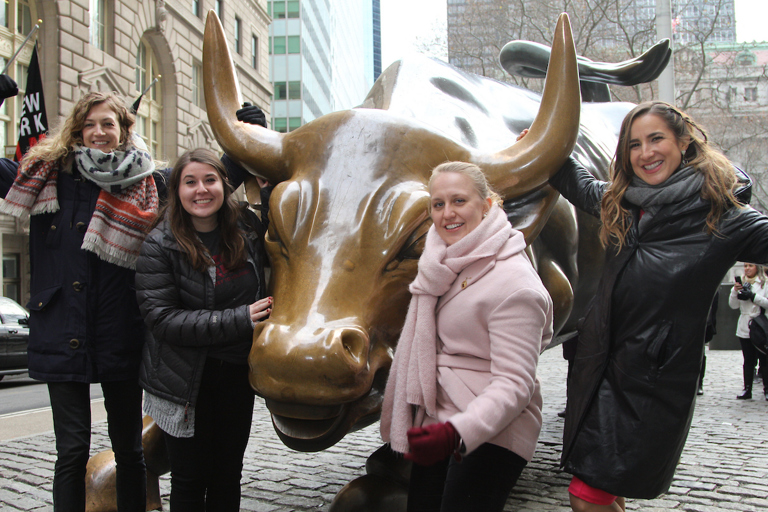 Four women pose with a statue of a golden bull in New York City.