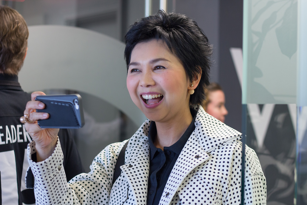 A stylish, smiling Asian woman takes a video selfie with her phone.