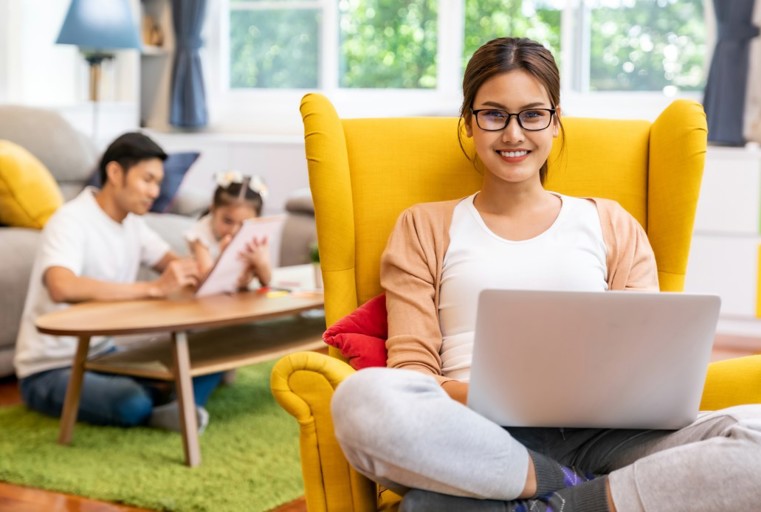A woman sits in a yellow chair working on a laptop while her husband and child play quietly nearby.