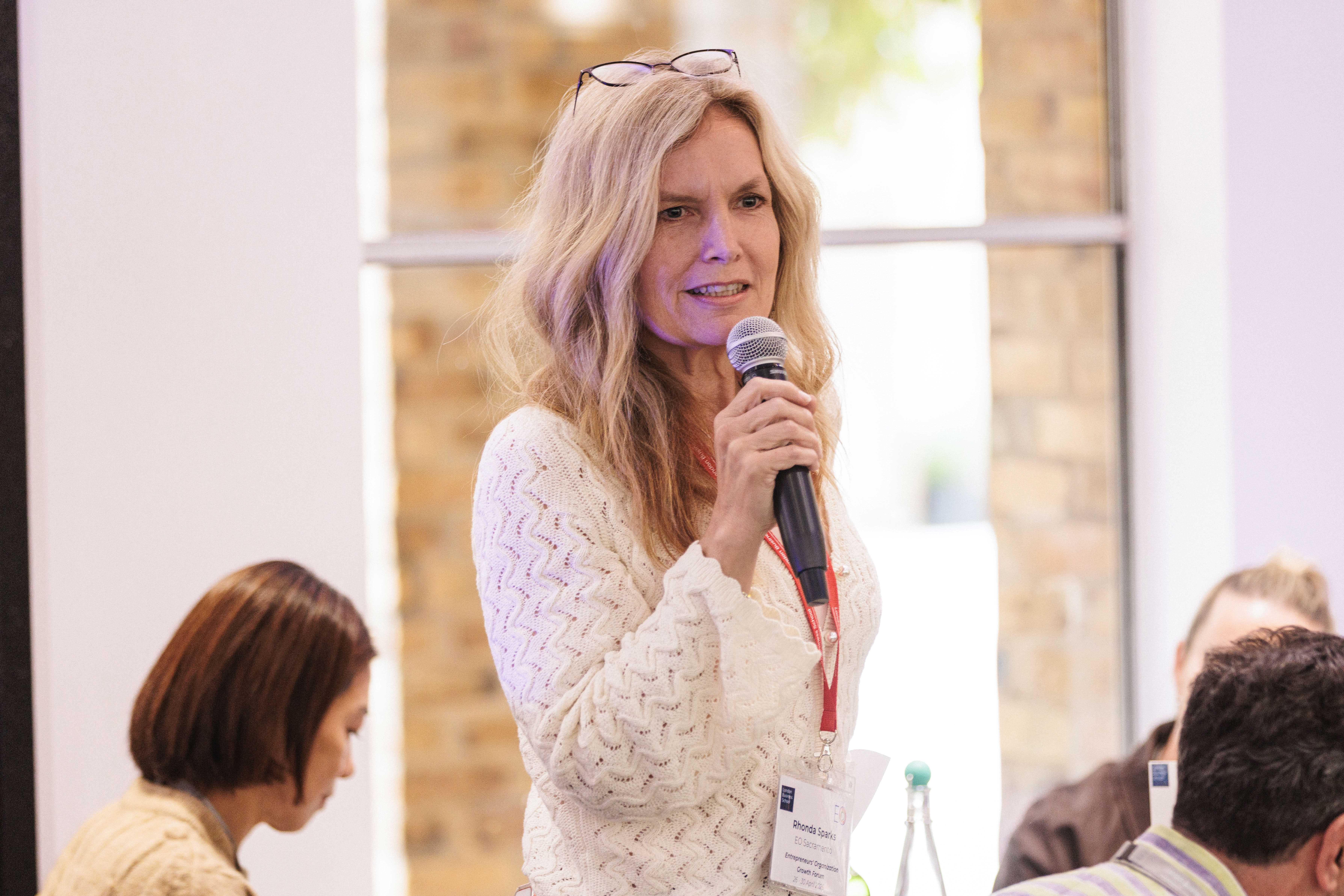 A professional woman speaks into a microphone in a classroom setting.