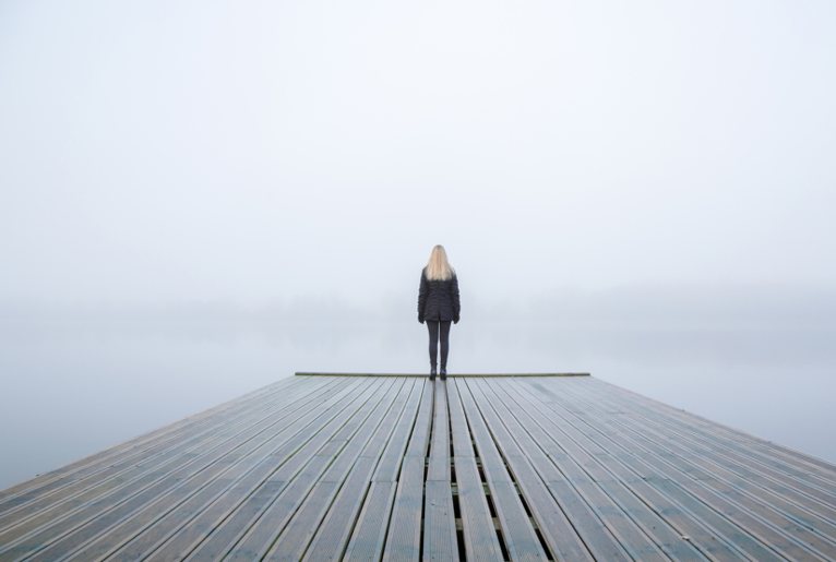 A lone woman stands at the end of a pier, looking out into foggy skies.