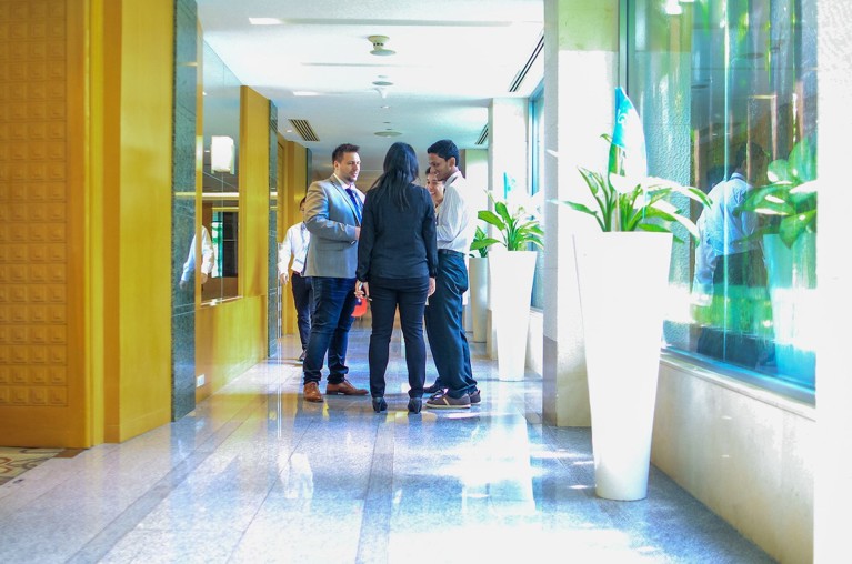 A trio of business persons conduct a discussion in a brightly lit hallway.