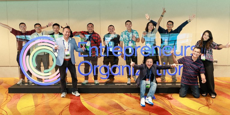A group of enthusiastic EO members pose around a large EO logo sign.