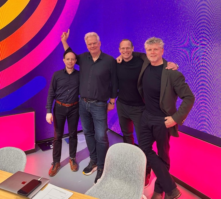 Four men stand in front of a wall featuring the EO logo.