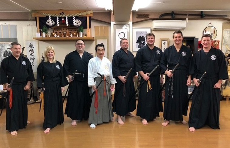 Eight men and women dressed in Samurai garb holding Samurai swords pose in a Tokyo dojo.