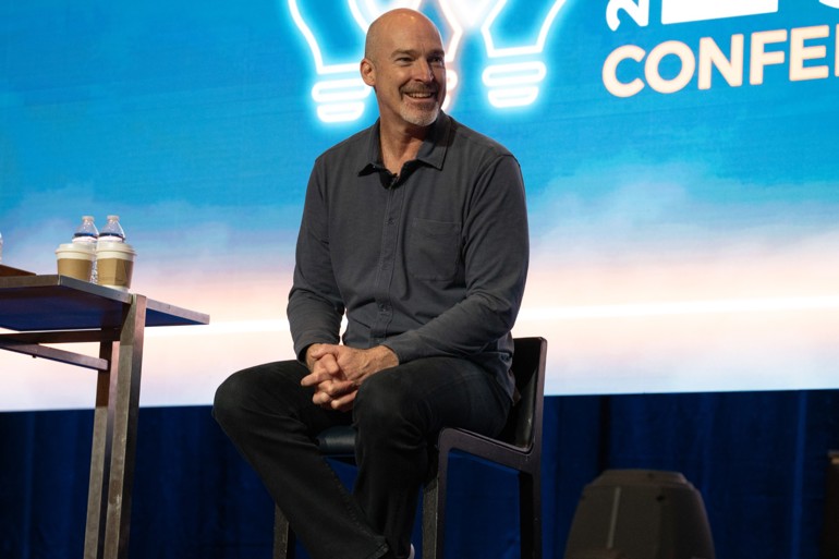 A man sits on a stage presenting to a room full of entrepreneurs.