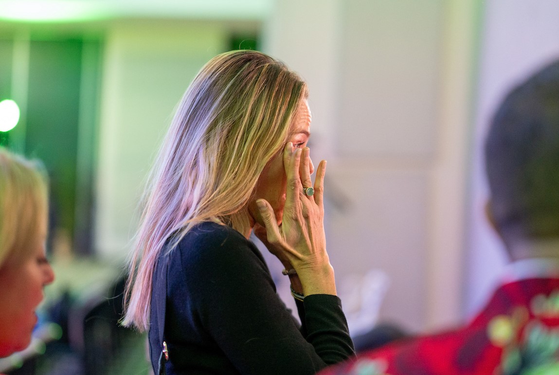 A woman in profile, dressed in business attire, wipes a tear from her eye,