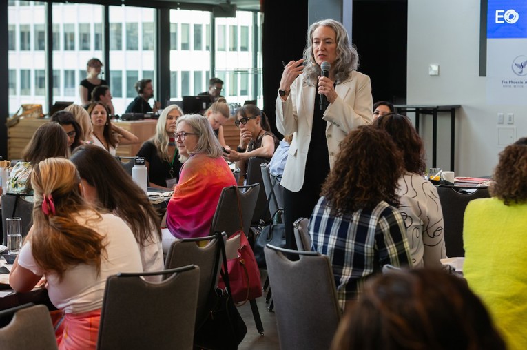 A woman with a microphone speaks amid a room full of women at lunch tables.