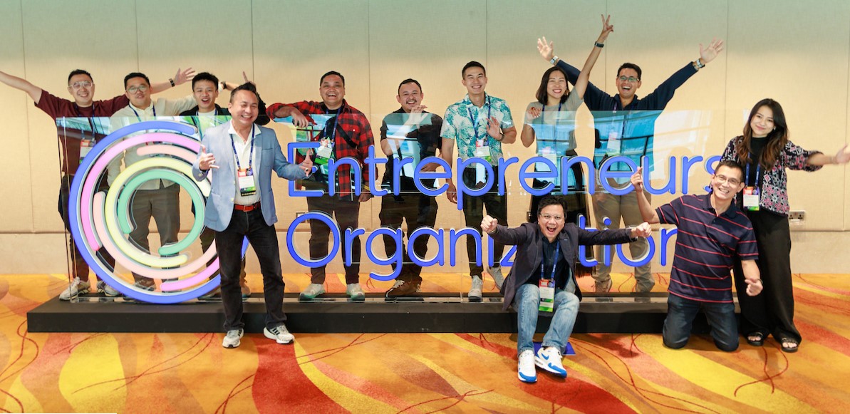 A group of 15 EO members pose with enthusiasm by an EO logo sign.