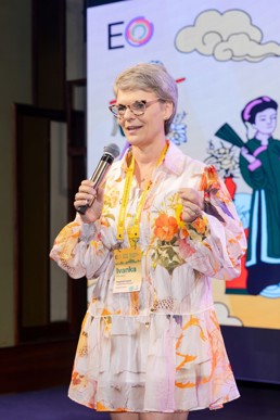 A woman in a floral dress holds a microphone as she makes a presentation.